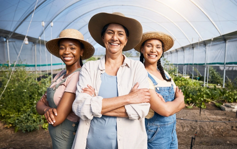 Three women working on a farm, standing together in a field with crops in the background. They are dressed in casual farm clothes and smiling at the camera.