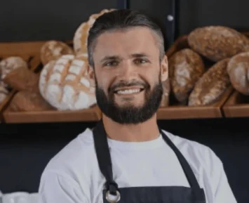 A man behind the counter in a bakery, serving a customer with a friendly smile. Freshly baked bread and pastries are displayed on shelves behind him