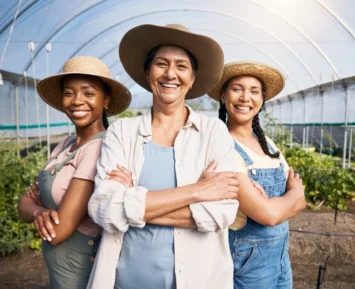 Three women working on a farm, standing together in a field with crops in the background. They are dressed in casual farm clothes and smiling at the camera.